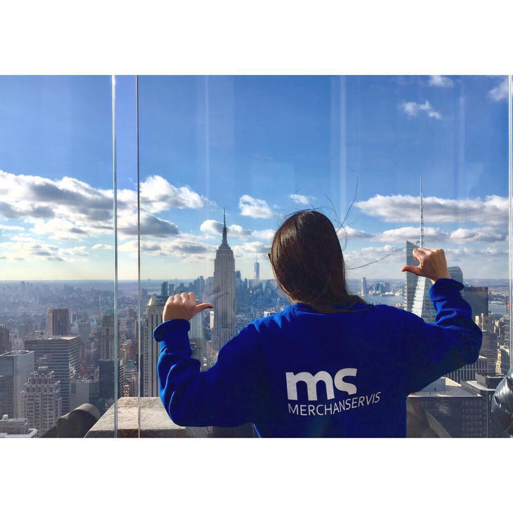 La imagen muestra a una chica con una camiseta de Merchanservis en lo alto de un edificio señalándose la camiseta con los dedos mientras mira la ciudad.