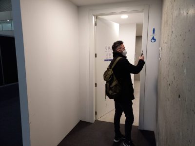 La imagen muestra una persona escaneando con un móvil lo que parece ser un QR al lado de la entrada de los baños.